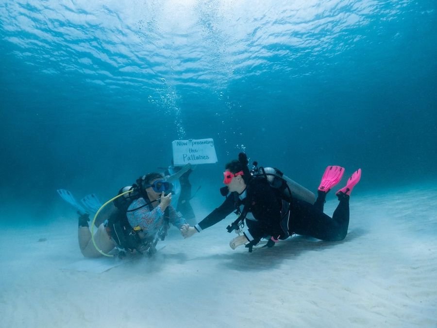 Mariya and Sam scuba diving on the sand holding a sign that says Now presenting the Pallaises