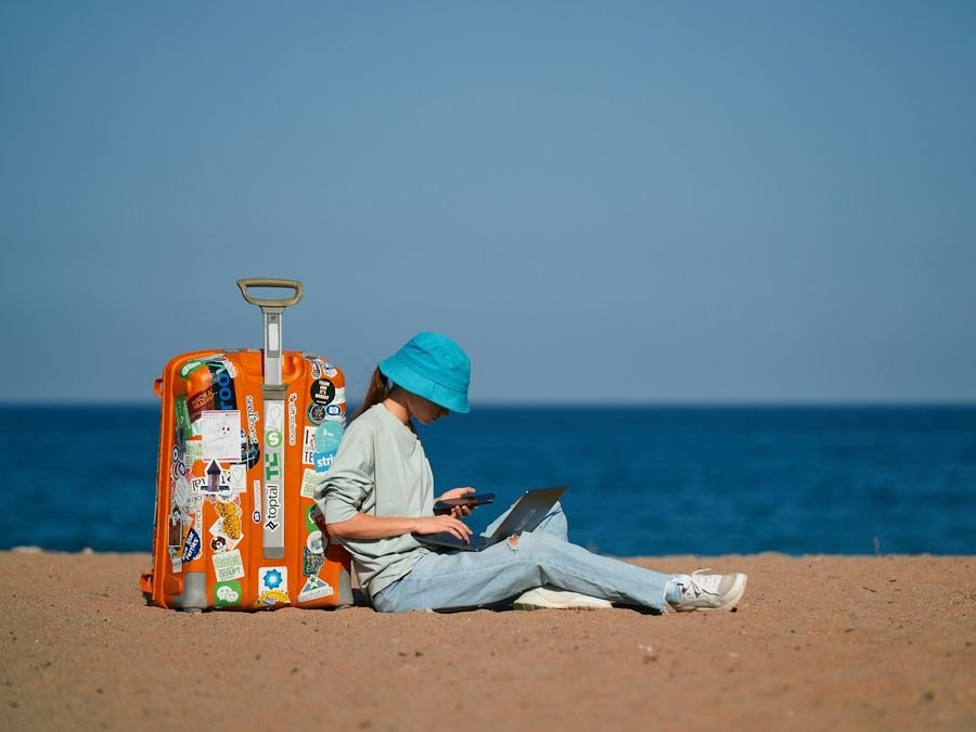 Woman working on a laptop on the beach beside a sticker-covered suitcase